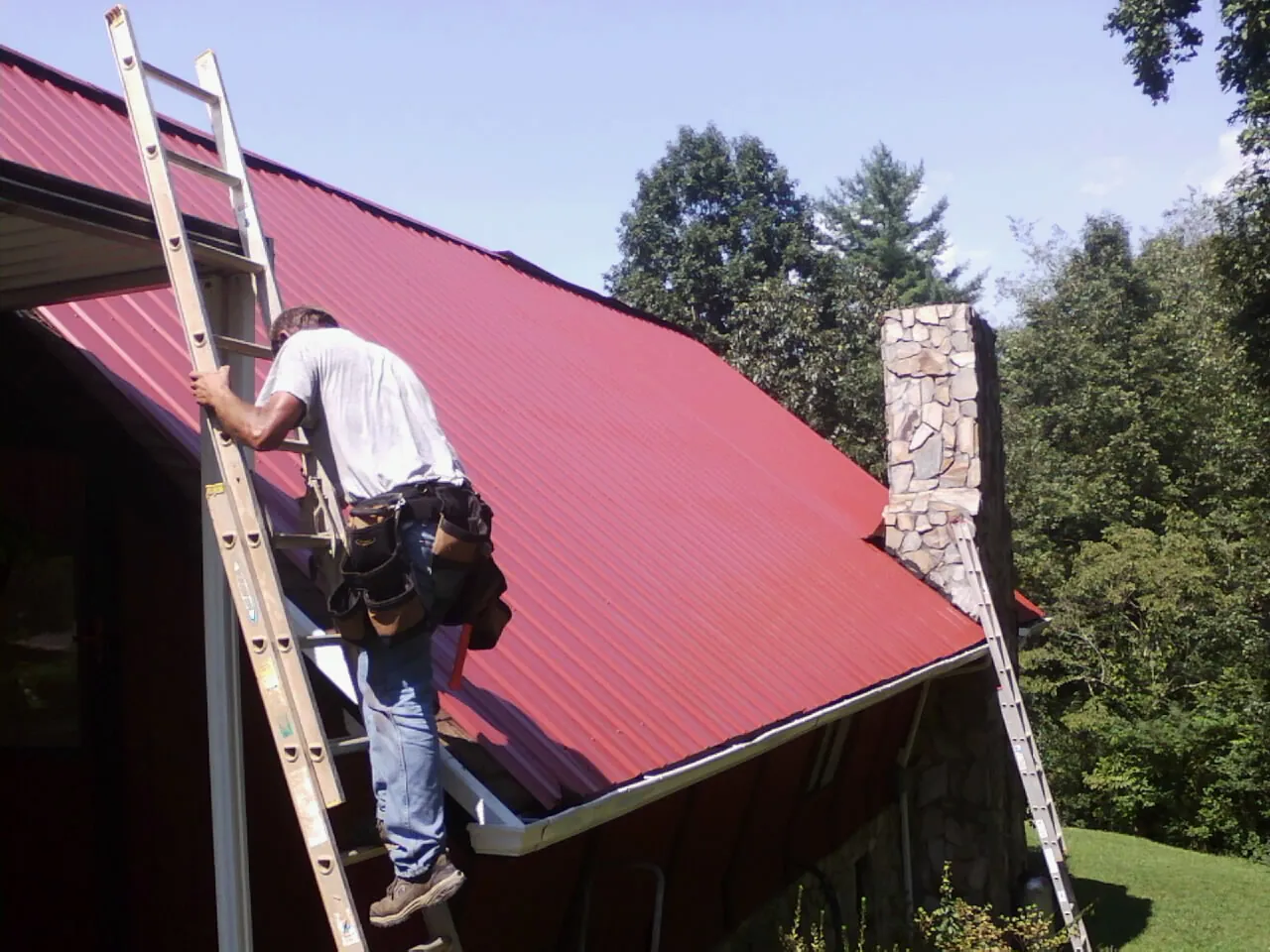 Roofer on a ladder beside a red metal roof and stone chimney — Soffit & Fascia Repair by Roofing Brothers in Westerlo